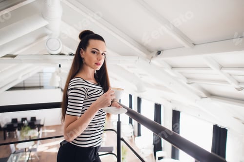 Visualização: Retrato de uma linda jovem morena tomando uma xícara de café em um café olhando para a câmera.