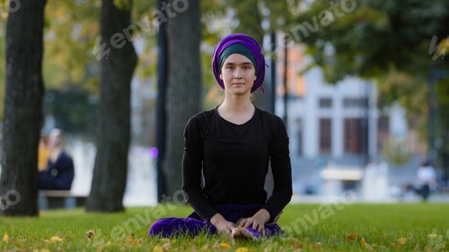 Preview: Young girl muslim woman in hijab sits on green grass in park takes leg by foot bends turns to side