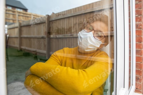 Preview: Woman wearing a face mask isolating at home and looking out of the window