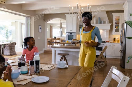 Preview: Smiling african american mother with breakfast walking towards daughter at dining table