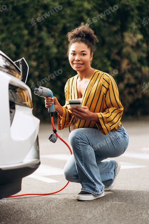 Preview: Smiling woman with phone holding electric car recharging at public parking car charging station