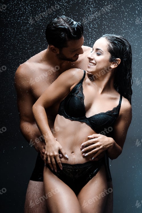 Preview: happy shirtless man hugging cheerful wet girl under raindrops on black