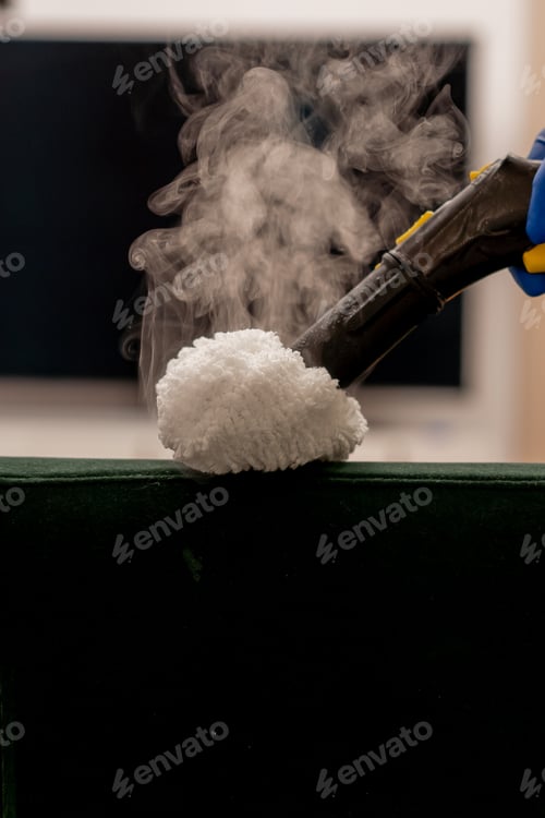 Preview: apartment cleaner in yellow apron does a steam cleaning of a green sofa with professional equipment