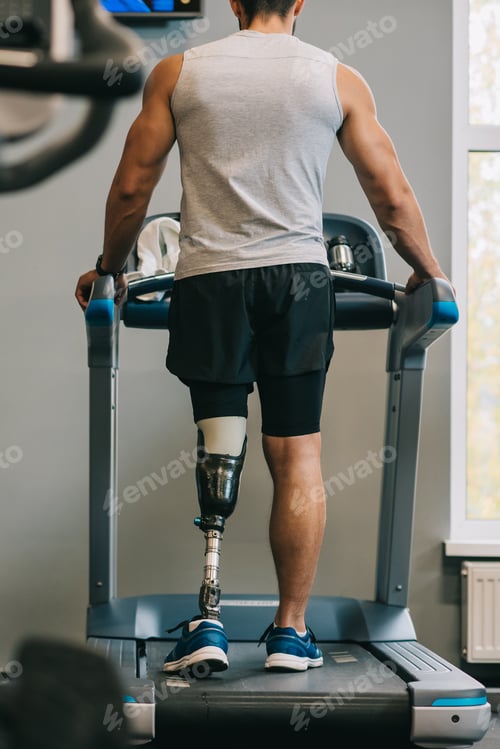 Preview: rear view of young sportsman with artificial leg walking on treadmill at gym