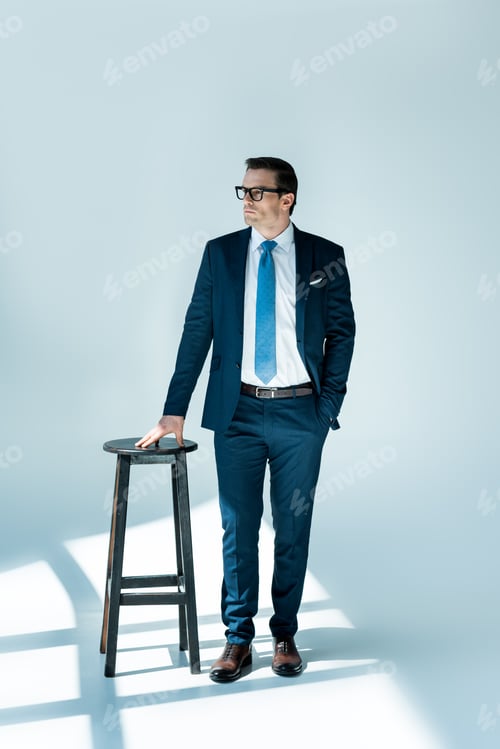 Preview: full length view of serious buisnessman in eyeglasses standing near stool and looking away on white
