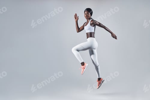 Preview: Full length of confident young African woman in sports clothing jumping against gray background