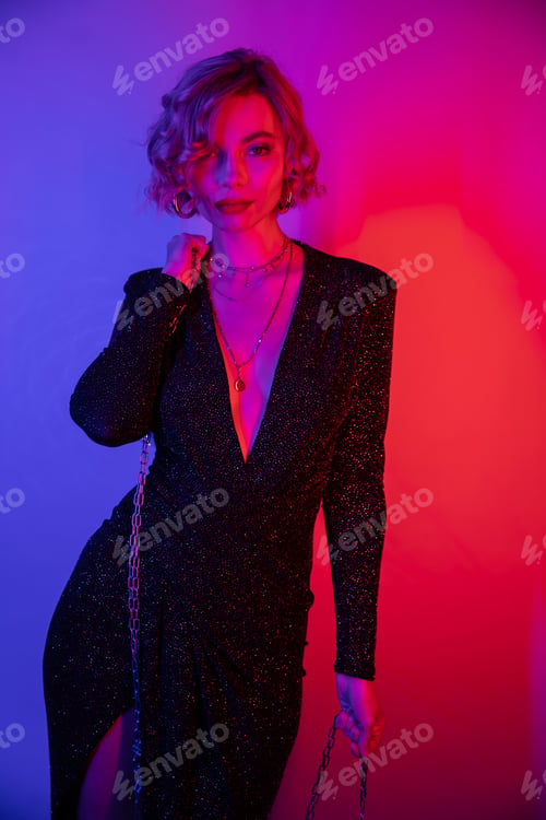 Preview: young woman with blonde hair standing in black tight dress and holding chain on purple and pink