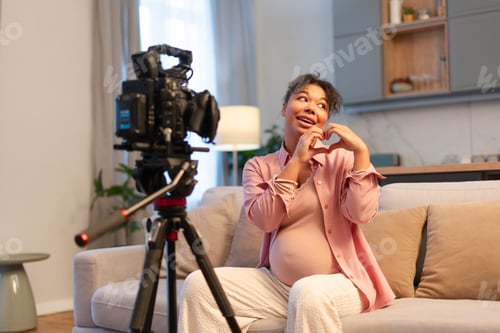 Preview: Positive beautiful African American pregnant woman showing heart with hands using camera on tripod