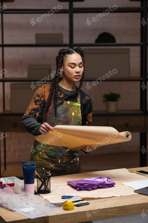 Preview: African american craftswoman holding craft paper near cloth in workshop