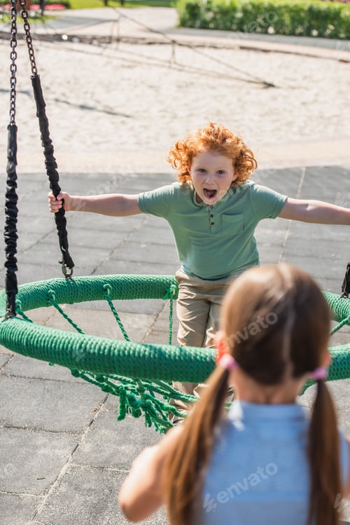 Preview: thrilled boy with open mouth having fun in hammock near blurred sister