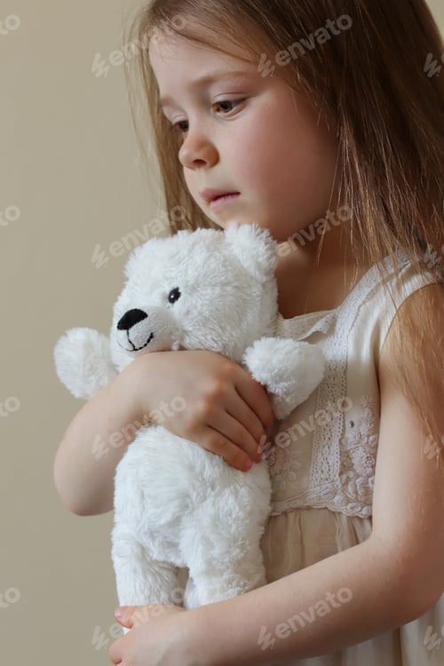 Preview: A little girl in a white dress holds a teddy bear in her hands