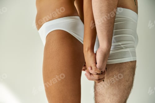 Preview: cropped view of young sexy multicultural couple posing back to back and holding their hands