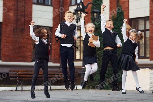 Preview: Group of kids in school uniform jumping and having fun outdoors together near education building
