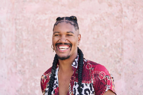 Preview: Young androgynous man with braids smiling and wearing shirt with animal print