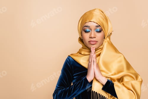 Preview: young muslim woman in hijab standing with praying hands isolated on beige