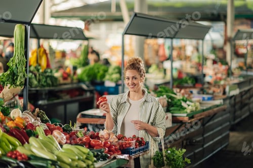 Preview: Happy buyer with tomato in hand standing near stall and smiling at camera