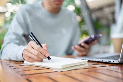 Preview: A man holding pen writing in notebook and other hand holding smartphone sitting at wooden table