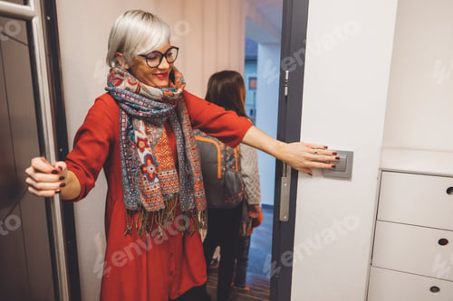 Preview: A woman with short blonde hair and glasses is opening a door, wearing a red dress and a colorful sca
