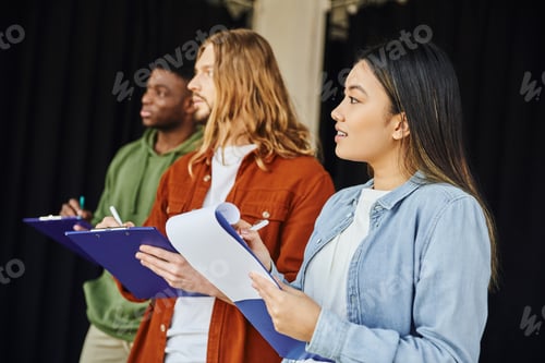 Preview: smiling asian woman writing on clipboard near multiethnic and stylish men during first aid seminar