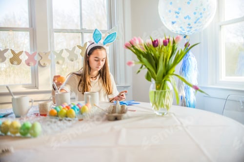 Preview: Teenage girl reading texts whilst dyeing hard boiled eggs for Easter at table