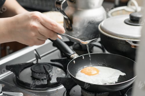 Preview: a girl's hand holding a spoon to pour the oil over the egg that is frying in a small frying pan.