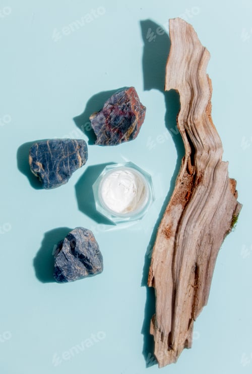 Preview: Cream jar next to stones and wood on blue background.