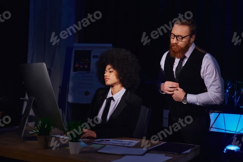 Preview: bearded businessman looking at pretty african american secretary typing on computer in night office