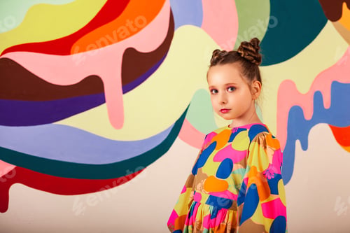 Preview: Serious sad little girl posing in colourful dress at multi colours wall, thought