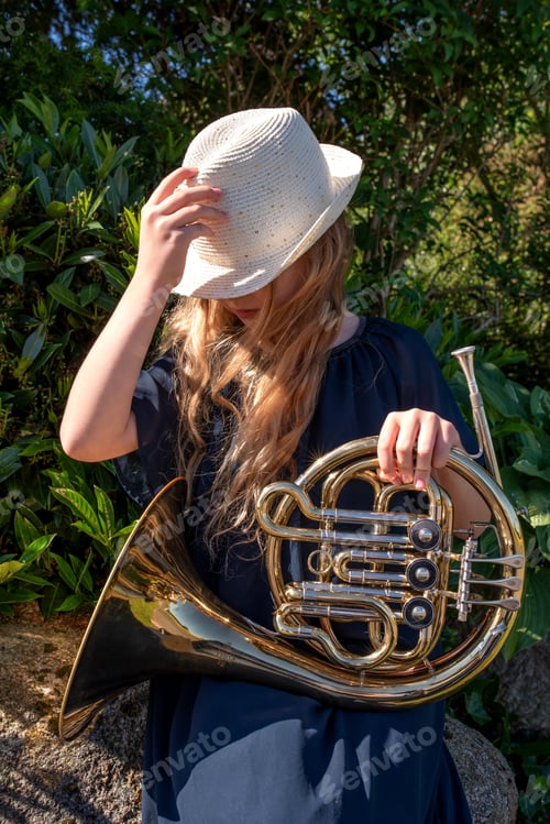 Preview: Blond girl in hat holds horn on her lap.