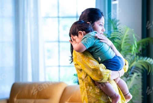 Preview: Indian Mom gently holding sleeping infant boy on shoulder, bonding quietly in bright modern home