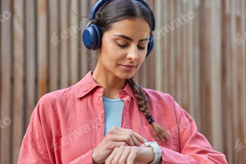 Preview: Horizontal shot of pretty brunette woman checks time on watch waits for someone wears stereo headpho