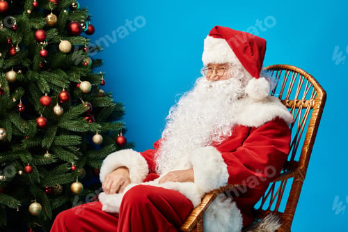 Preview: happy Santa Claus with beard and eyeglasses sitting in rocking chair near Christmas tree on blue