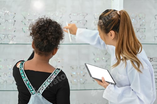 Preview: Smiling female optician helping child girl choosing the most appropriate eyeglasses.