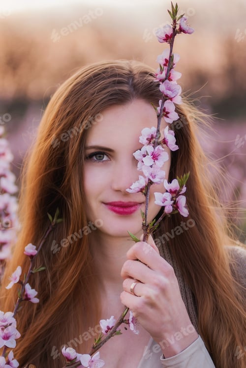 Preview: Close-up portrait of millennial woman holding blooming pink sakura branch near face. Natural beauty