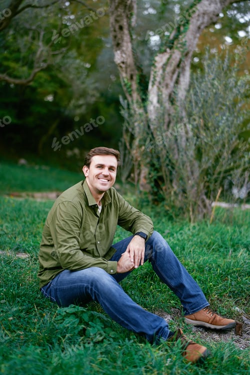 Preview: Smiling young man sitting on green grass in the park