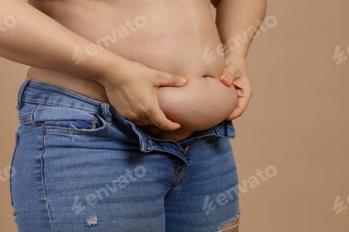 Preview: Woman holding big belly with hands showing fat in blue jeans on beige background. Sudden weight gain