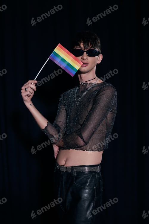 Preview: Trendy homosexual young man in sunglasses and shiny top biting lgbt flag and posing during pride