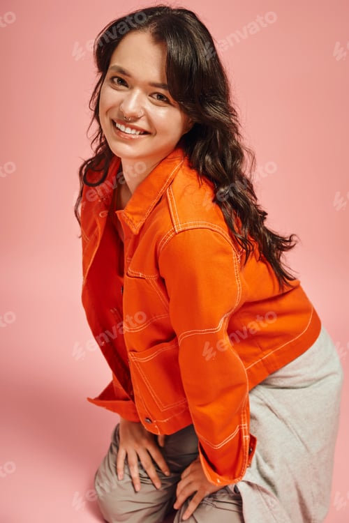 Preview: Stylish young woman poses gracefully in vibrant orange attire against a soft pink background