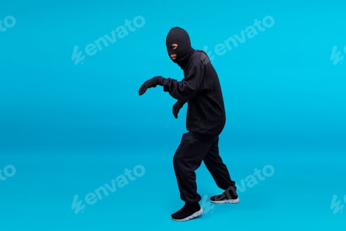 Preview: Full Length Of Burglar In Black Clothes Wearing Mask Walking Silently Isolated On Blue Background