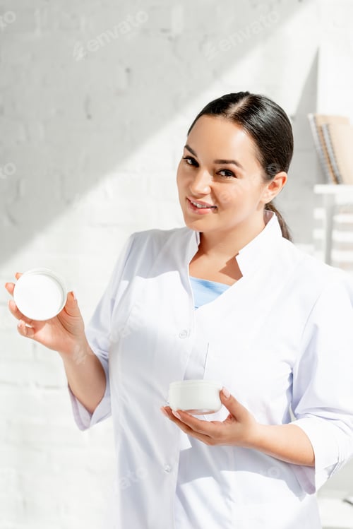 Preview: smiling dermatologist holding cosmetic cream and looking at camera in clinic