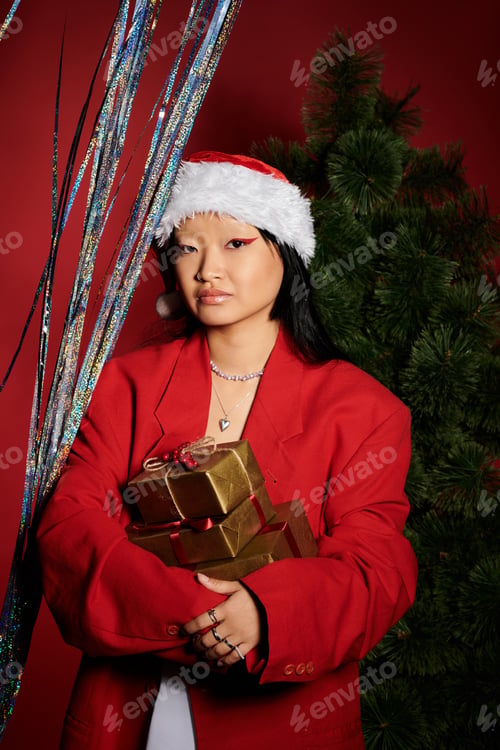 Preview: A stylish Asian woman in a red blazer and Santa hat holds gift boxes by a festive tree backdrop