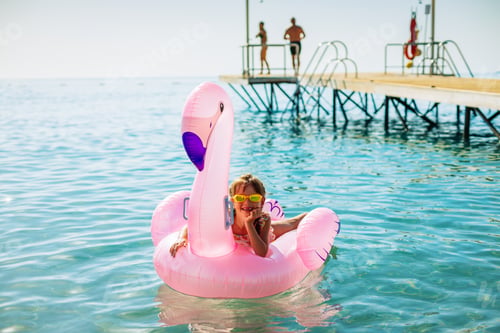 Preview: Happy laughing little girl in sunglasses lying on a pink flamingo inflatable float in the sea