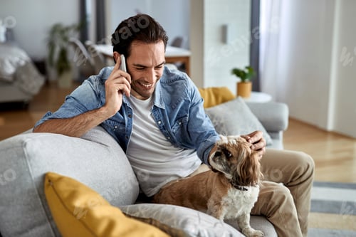 Preview: Young happy man talking on the phone while relaxing with his dog at home.