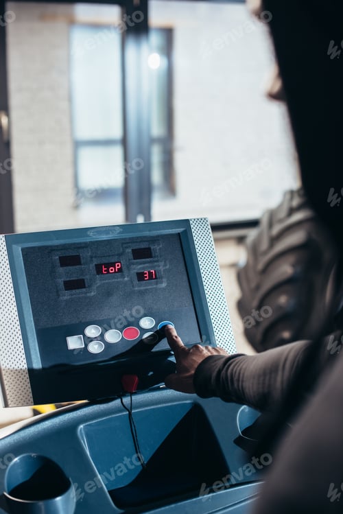 Preview: cropped image of sportsman in hoodie pushing button on treadmill in gym