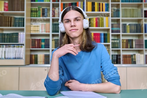 Preview: Teenage college student in headphones looking at webcam, sitting at desk in library