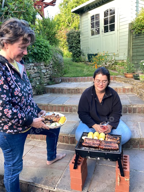 Preview: Two women in garden, cooking chicken, sausages and sweetcorn on a portable barbecue