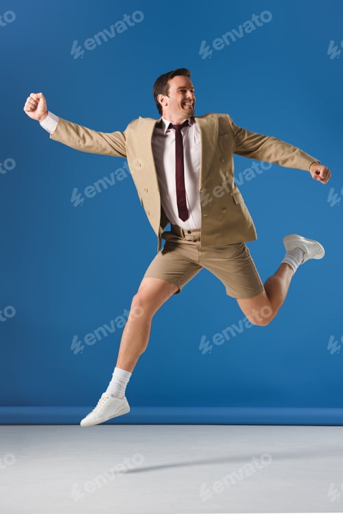 Preview: excited man in shorts and suit jacket jumping and looking away on blue