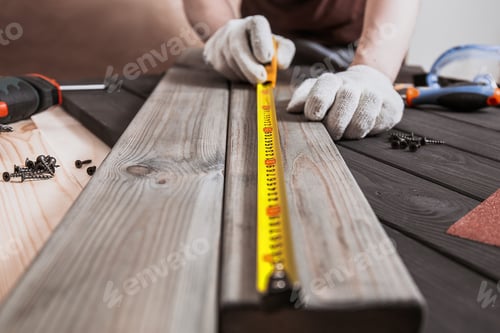 Preview: Male carpenter in gloves making measurements on the wooden plank by yellow measure tape