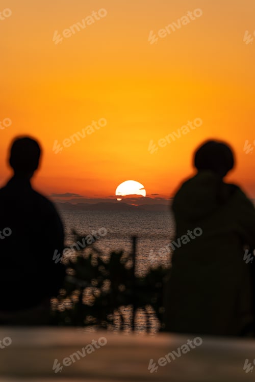 Preview: Silhouettes of two people watching a bright orange sunset over the sea
