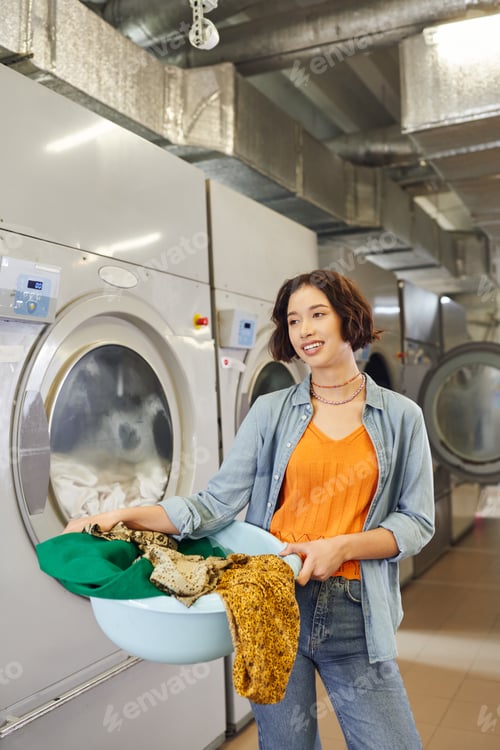 Preview: cheerful young asian woman holding basin with clothes near washing machines in public laundry